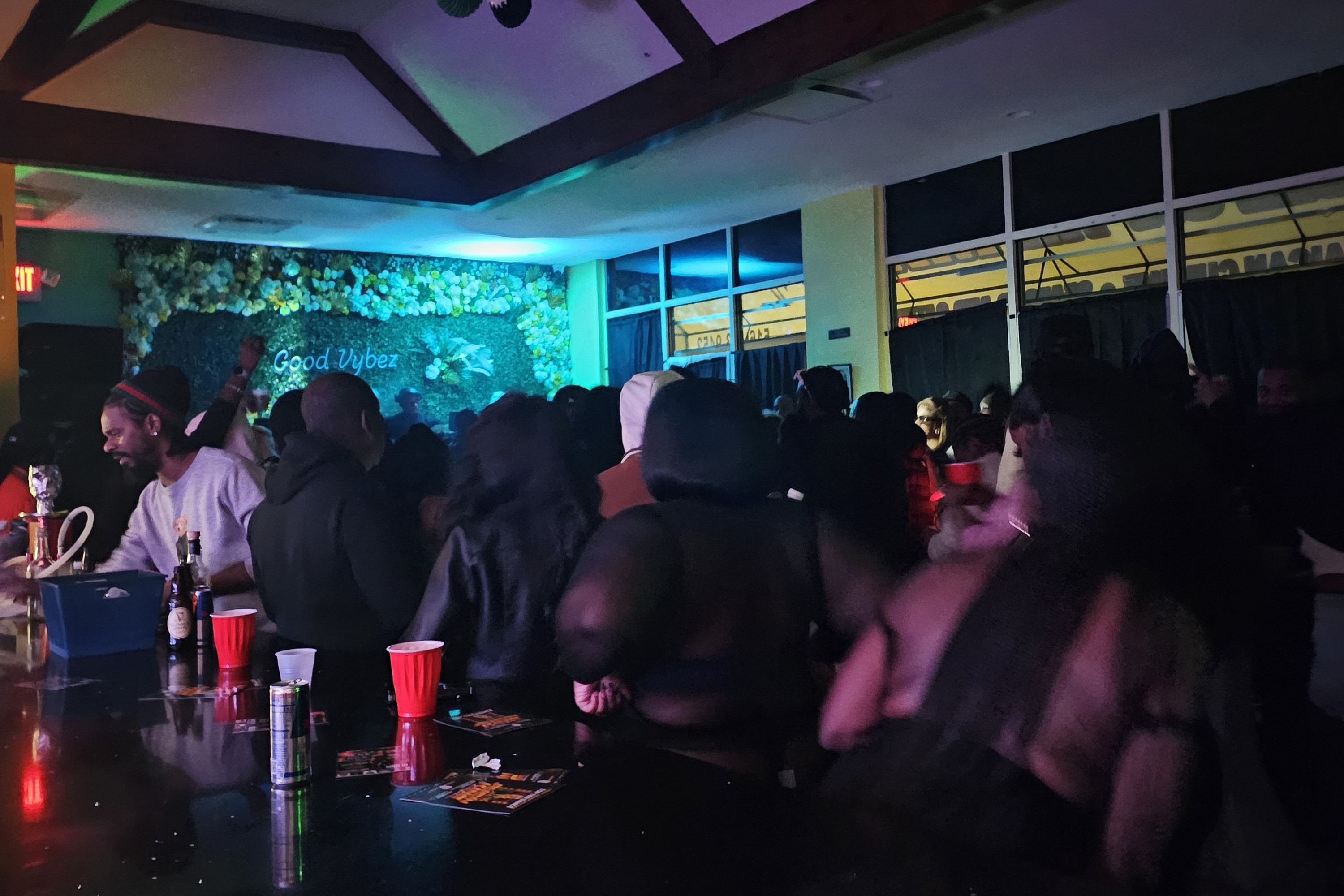 Private partygoers stand with their backs to the bar, and face the neon blue Good Vybez sign over floral backdrop above the stage.