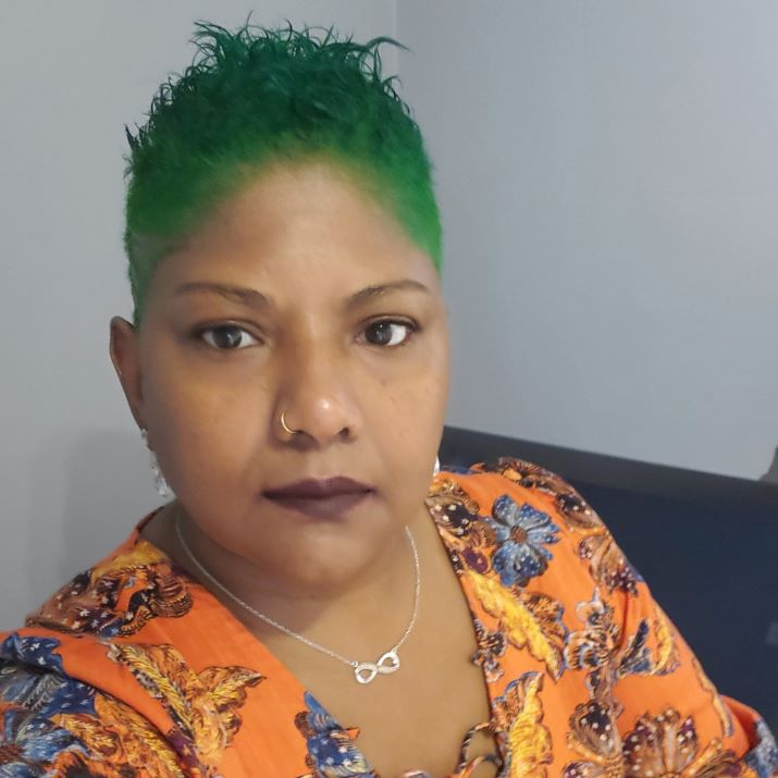 Meloney, an indo-Jamaican woman with a bright green spiked low haircut, a gold hoop nose ring, silver necklace, and dark lipstick wears a bright orange top with blue floral accents.
